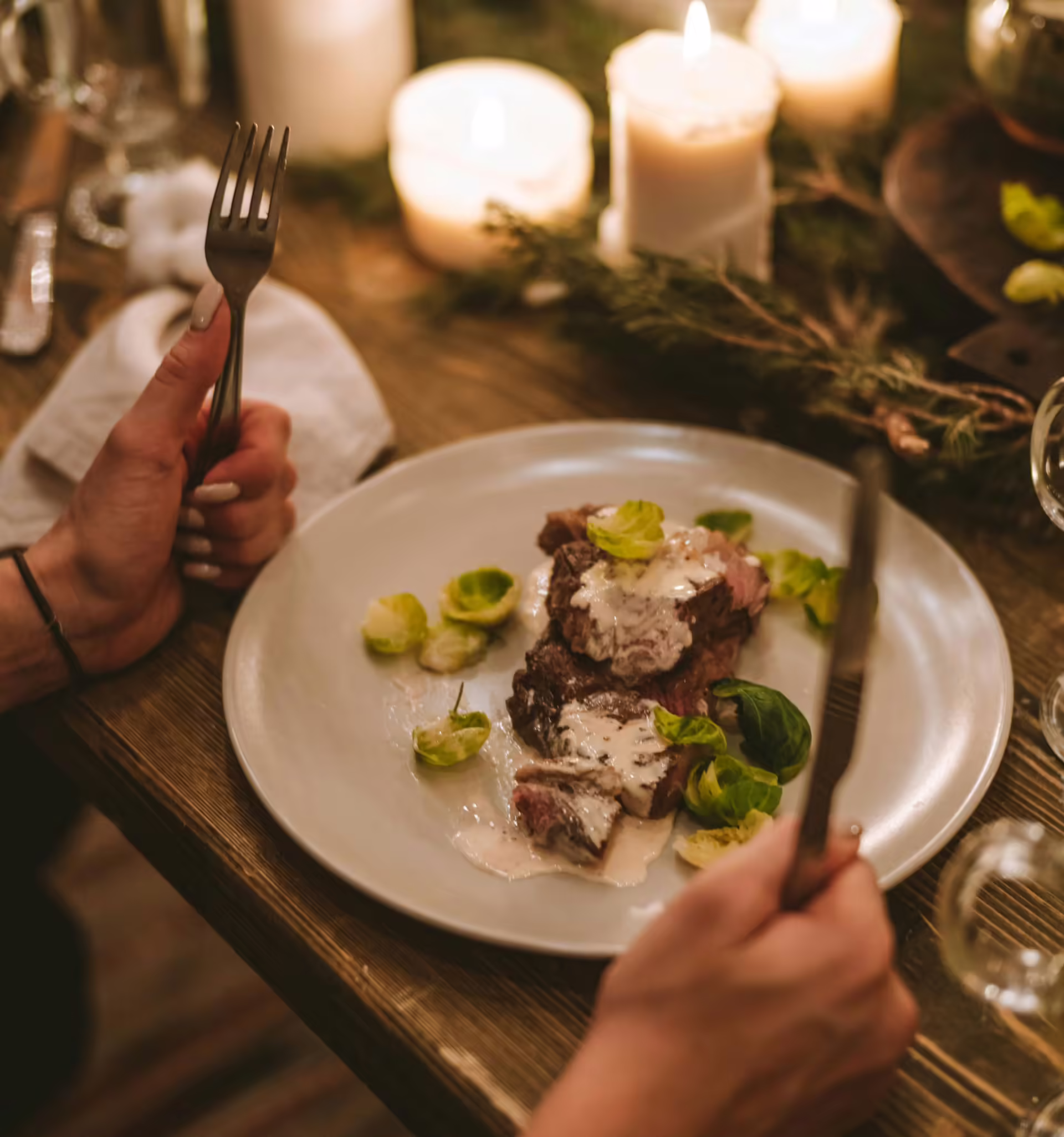 A person holding a fork and knife, preparing to enjoy a steak dinner with brussels sprouts, surrounded by candlelight.