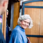 An older woman with curly gray hair smiling while walking through a doorway.