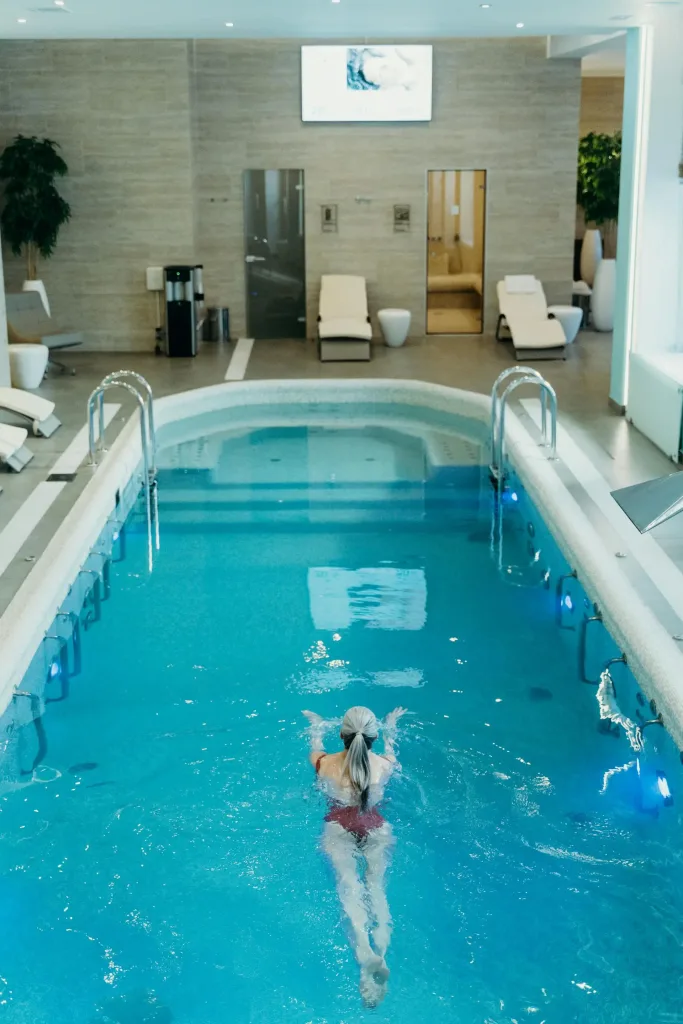 A senior woman swimming in a serene indoor pool surrounded by modern spa-like amenities, including comfortable lounge chairs and a calming atmosphere.