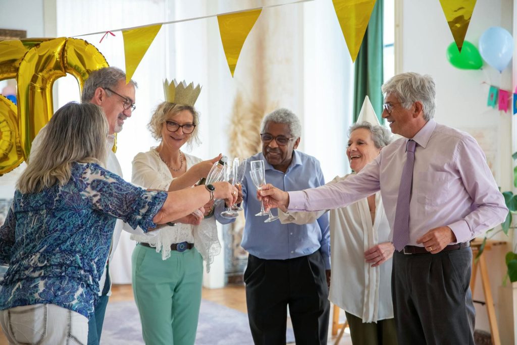 A group of cheerful seniors celebrating together with champagne, wearing festive attire and surrounded by colorful decorations and balloons.
