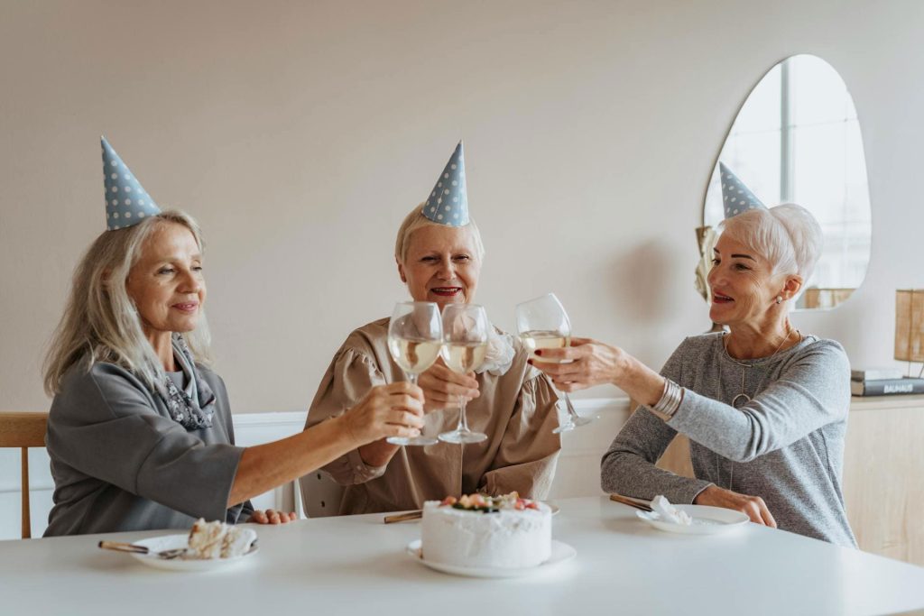 Three senior women wearing party hats clinking glasses of white wine at a table with a celebratory cake, enjoying a cheerful moment together.