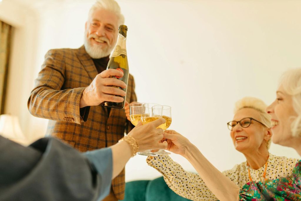 A joyful gathering with a smiling man in a plaid blazer pouring champagne into glasses held by three women, celebrating with laughter and cheer.
