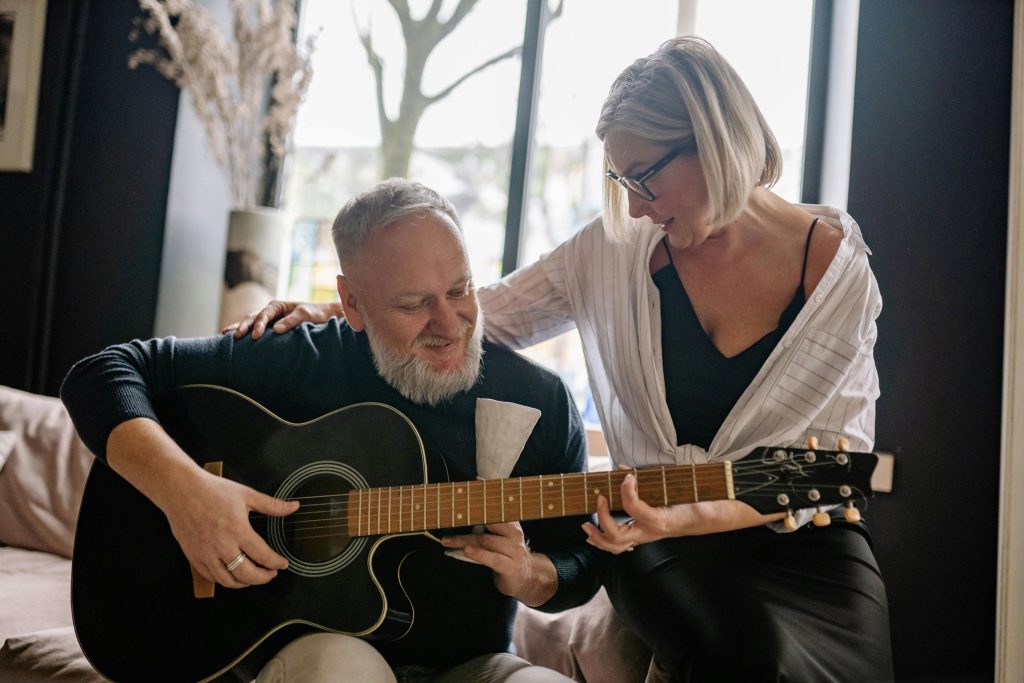 A smiling man plays a guitar while a woman lovingly places her hand on his shoulder, enjoying a moment of music and connection together.